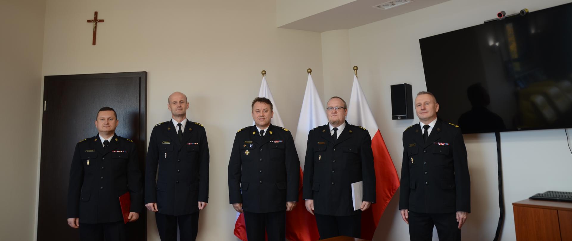 W pomieszczeniu stoi pięciu mężczyzn ubranych w stroje wyjściowe PSP. Każdy mężczyzna patrzy wprost. Za mężczyznami są drzwi, trzy flagi Polski i telewizor.