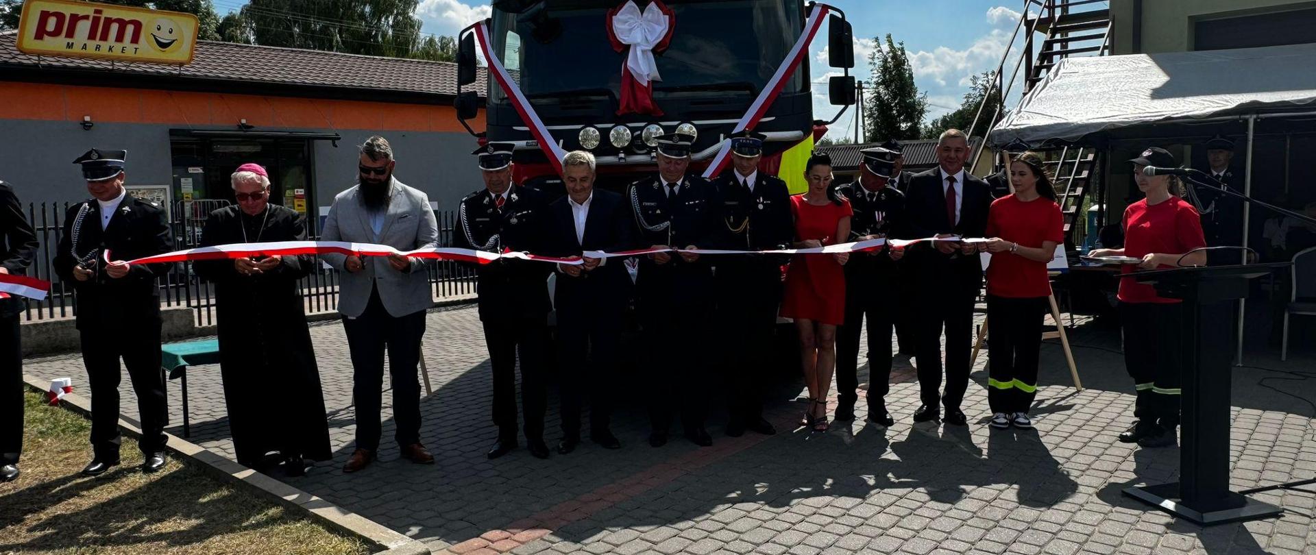Zaproszeni goście oraz członkowie zarządu OSP przecinają wstęgę przed nowym samochodem gaśniczym