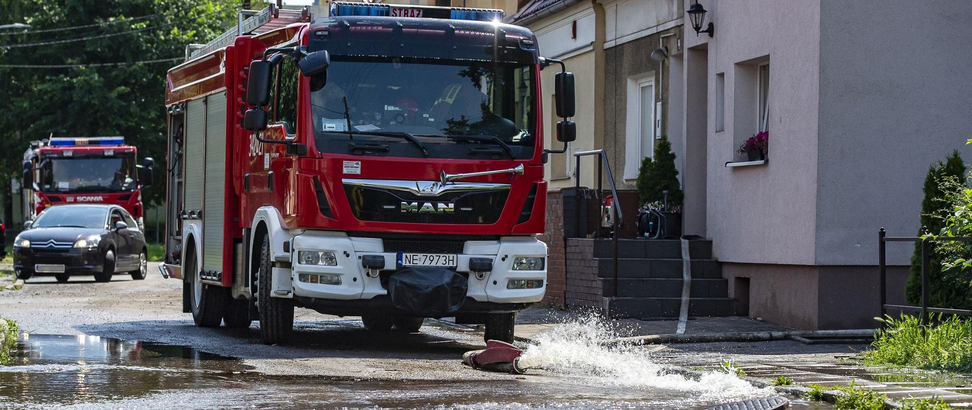 Na jezdni stoi pojazd strażacki, przed nim rozwinięty jest wąż strażacki z którego wylewa się na ulicę woda, wypompowywana z pobliskiego budynku.