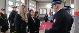 Komendant Miejski wręcza listy gratulacyjne pracownikom cywilnym Komendy, W tle poczet sztandarowy i inni strażacy zgromadzeni na zbiórce.
