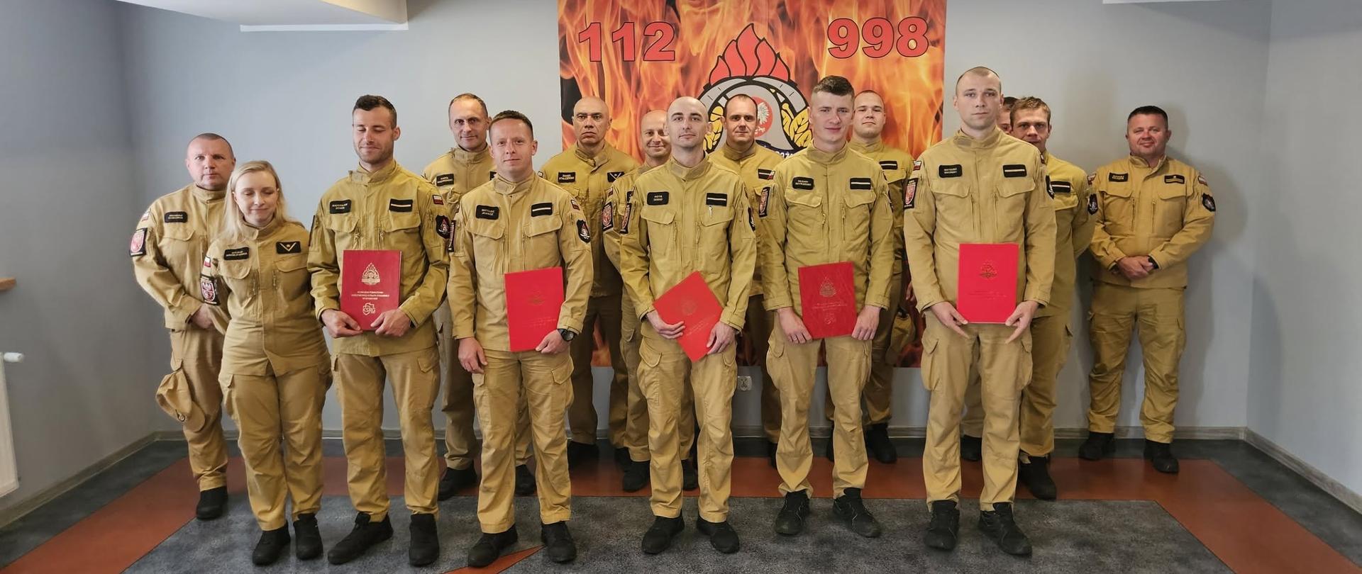Grupa strażaków Państwowej Straży Pożarnej w umundurowaniu koszarowym pozuje do wspólnego zdjęcia. Kilka osób trzyma czerwone teczki z aktami awansów. W tle widoczny jest emblemat straży pożarnej oraz numery alarmowe 112 i 998.