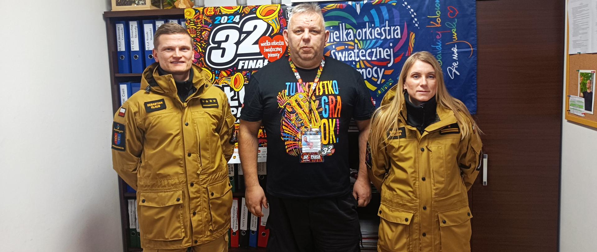 Do zdjęcia na stojąco pozuje 2 strażaków w umundurowaniu służbowym oraz szef sztabu WOŚP rejon Ostrowiec Św. Zdjęcie wykonane w siedzibie sztabu WOŚP teren Ostrowiec Św. W tle plakat WOŚP 32 Finał.