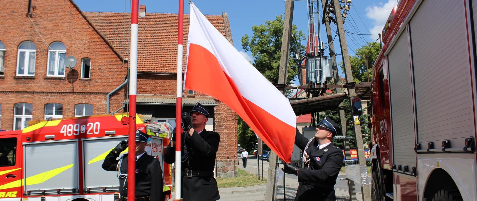 Strażacy uroczyście wciągają flagę państwową na maszt. Po bokach stoją wozy strażackie.