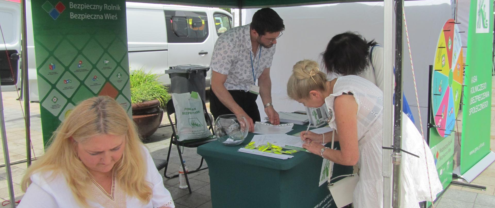 Na fotografii widać stoisko KRUS w pełnej okazałości. Stolik przyozdobiony obrusem z logiem Kasy idealnie współgra kolorystycznie z banerami oraz kołem prewencyjnym. Pracownik PT KRUS w Ciechanowie Mateusz Krakowski obsługuje uczestników gry prewencyjnej, wpisujących się na listę. Na pierwszym planie kobieta w skupieniu wypełnia test w ramach gry.