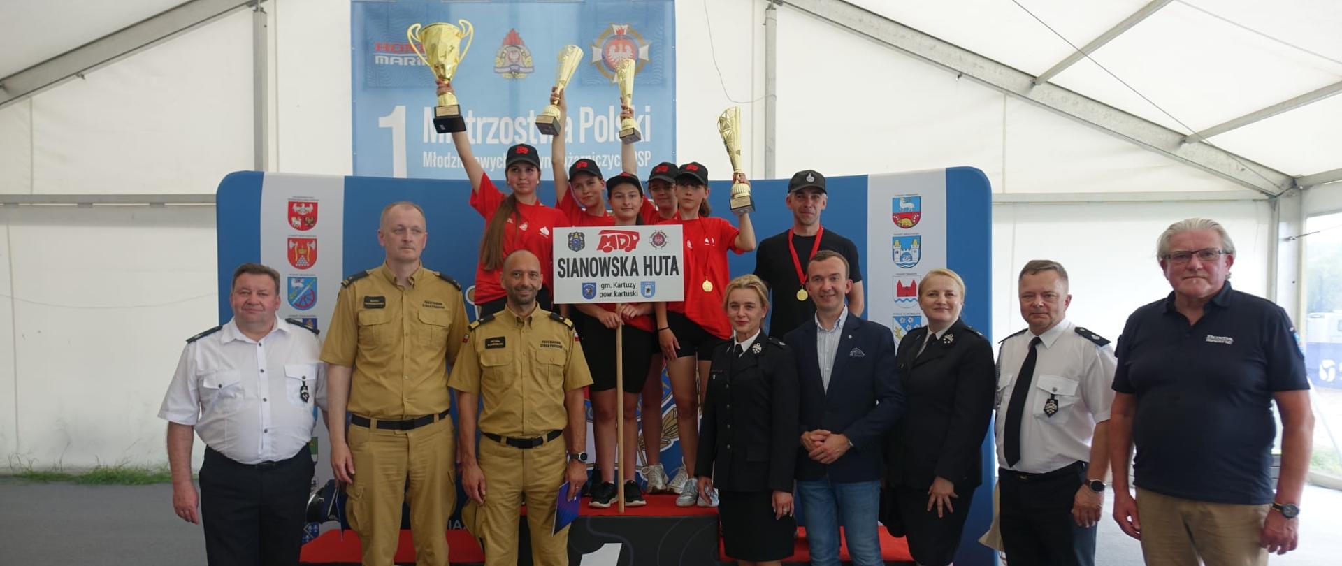 Zdjęcie przedstawia drużynę MDP Sianowska Huta stojącą na najwyższym stopniu podium. Obok nich pozują do zdjęcia organizatorzy zawodów oraz przedstawiciele straży pożarnej