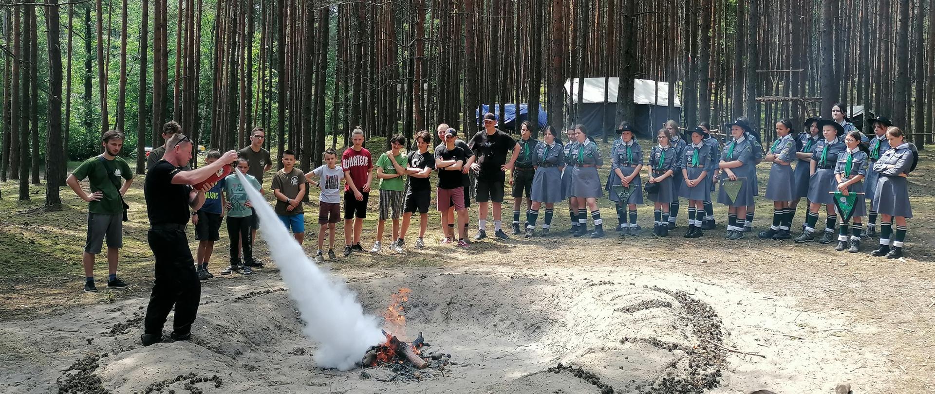 Fotografia przedstawia ujęcie miejsca ogniskowego w lesie. Pośrodku kadru w dole płonie ognisko. Dookoła ogniska grunt w promieniu dwóch metrów jest oczyszczony do warstwy mineralnej. Dookoła ogniska stoją harcerze i harcerki. Po lewej stronie kadru strażak w mundurze dowódczo-sztabowym podaje prąd proszku z gaśnicy na ognisko, harcerze przyglądają się. Dookoła rosną drzewa. Jest dzień.
