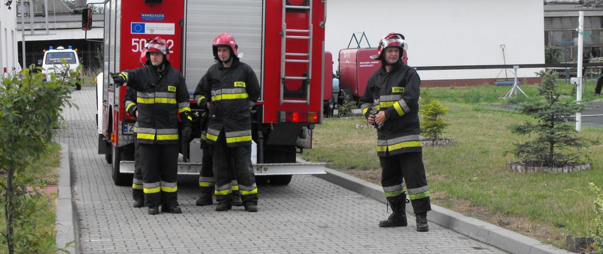 Zdjęcie przedstawia strażaków w ubraniach specjalnych przed samochodem pożarniczym