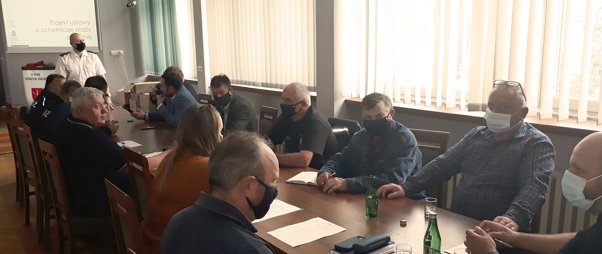 Na zdjęciu widać komendanta powiatowego PSP w Ustrzykach Dolnych ubranego w białą koszulę stojącego przy ekranie. Przed komendantem stoi stół przy którym siedzi dwanaście osób w ubraniach cywilnych. Są to strażacy OSP. Wszyscy mają na twarzach maseczki. 