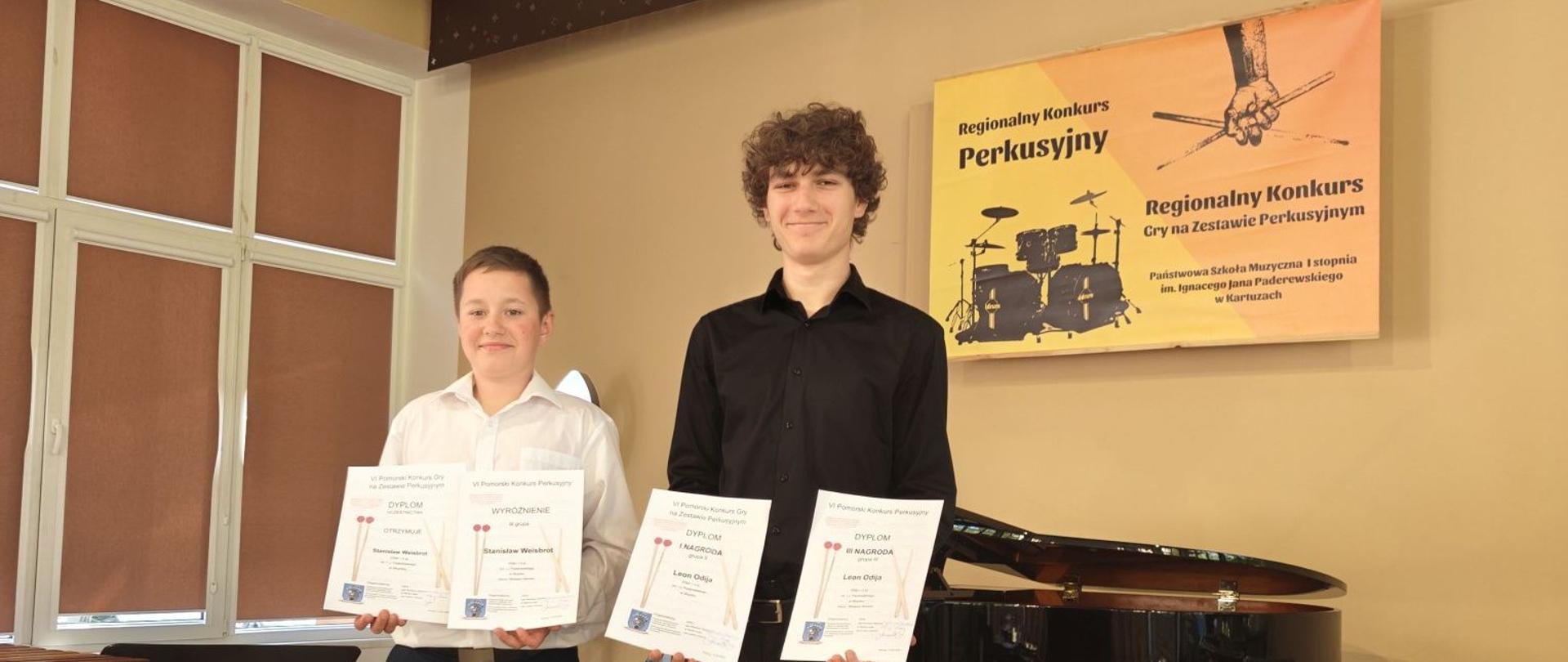 Zdjęcie kolorowe. Dwóch chłopców stoi w pomieszczeniu. Chłopiec po lewej stronie ma na sobie białą koszulę i ciemne spodnie. Chłopiec po prawej ma na sobie czarną koszulę i czarne spodnie. Obaj trzymają po dwa dyplomy. Za nimi widoczny jest fortepian i instrumenty perkusyjne. Na ścianie wisi plakat z napisem "Regionalny Konkurs Perkusyjny".