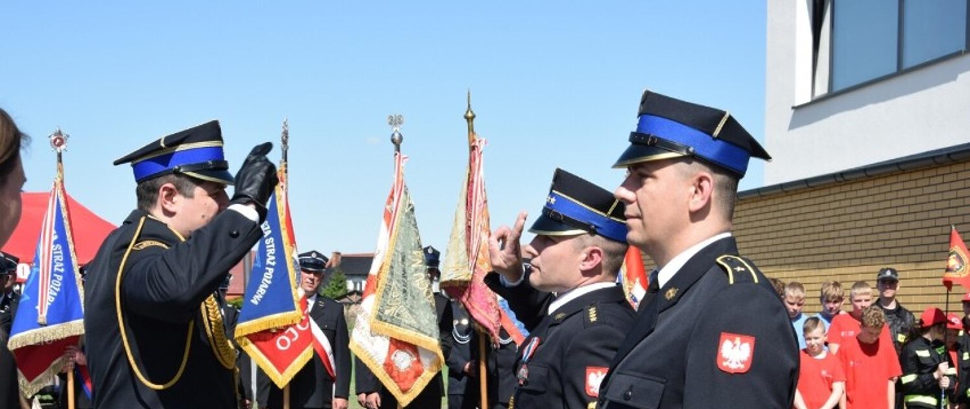 strażak salutuje innemu strażakowi który stoi na przeciwko, w tle poczty sztandarowe