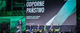 Minister Tomasz Siemoniak przy mównicy, w tle paneliści i duży ekran z nazwiskami panelistów i napisem Odporne państwo 
