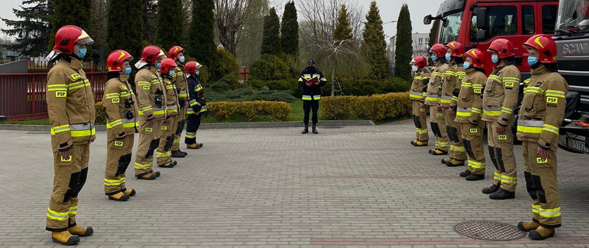 Zdjęcie przedstawia strażaków w umundurowaniu bojowym z założonymi maseczkami ochronnymi zakrywającymi usta i nos. Strażacy podczas uroczystej zmiany służby stoją w dwóch szeregach naprzeciwko siebie. Po prawej stronie znajdują się dwa samochody specjalne. Na drugim planie widnieje strażak w ubraniu dowódczo-sztabowym, który odczytuje rozkaz dzienny.