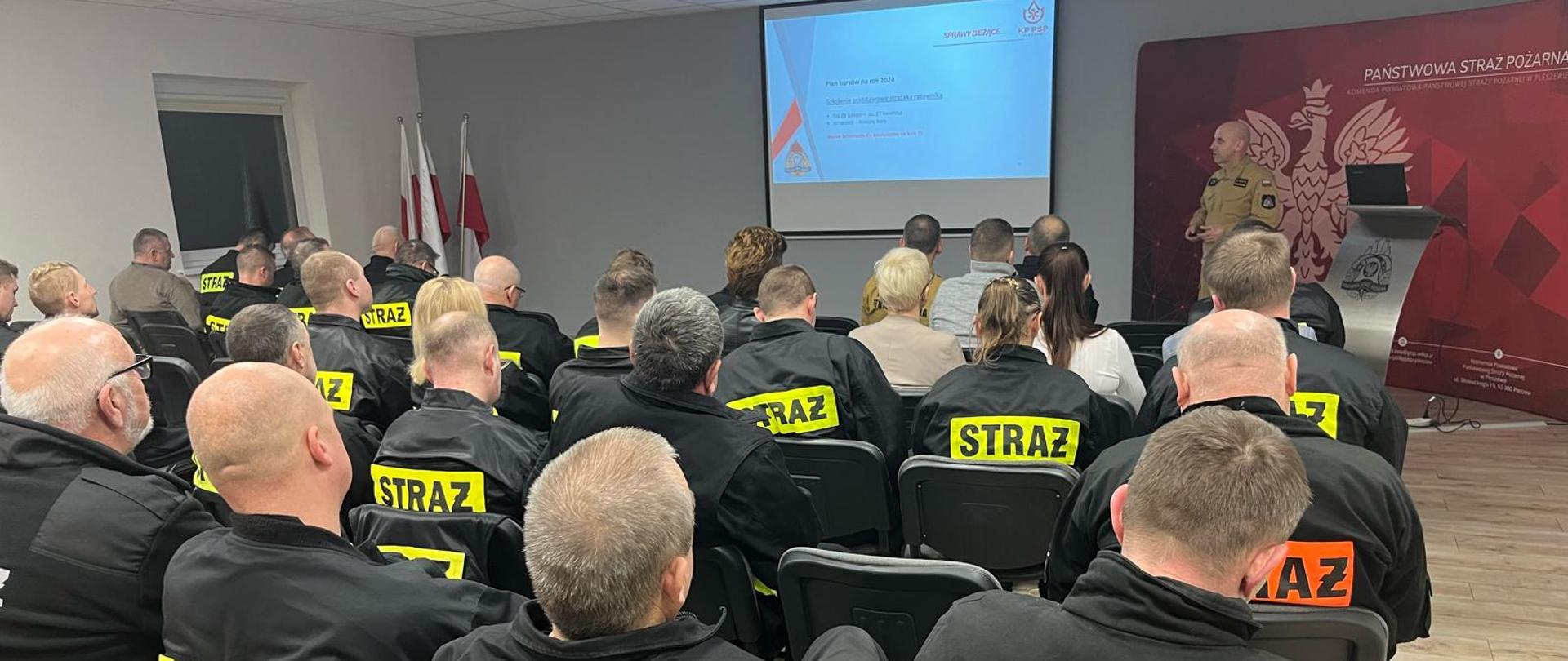 Na oświetlonej sali za mównicą stoi mężczyzna w mundurze państwowej straży pożarnej przemawiając do siedzących osób, za mężczyzną widać czerwony baner Państwowej Straży Pożarnej w Pleszewie