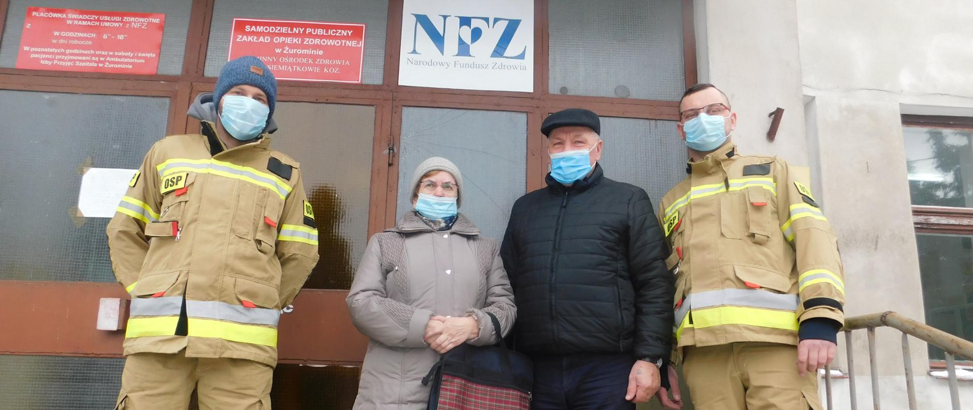 Czwórka ludzi stojących przed drzwiami budynku zakładu opieki zdrowotnej w maseczkach. Wśród ludzi po zewnętrznych stronach stoi dwóch strażaków w żółtych ubraniach specjalnych. Pomiędzy nimi stoi para starszych ludzi kobieta i mężczyzna.