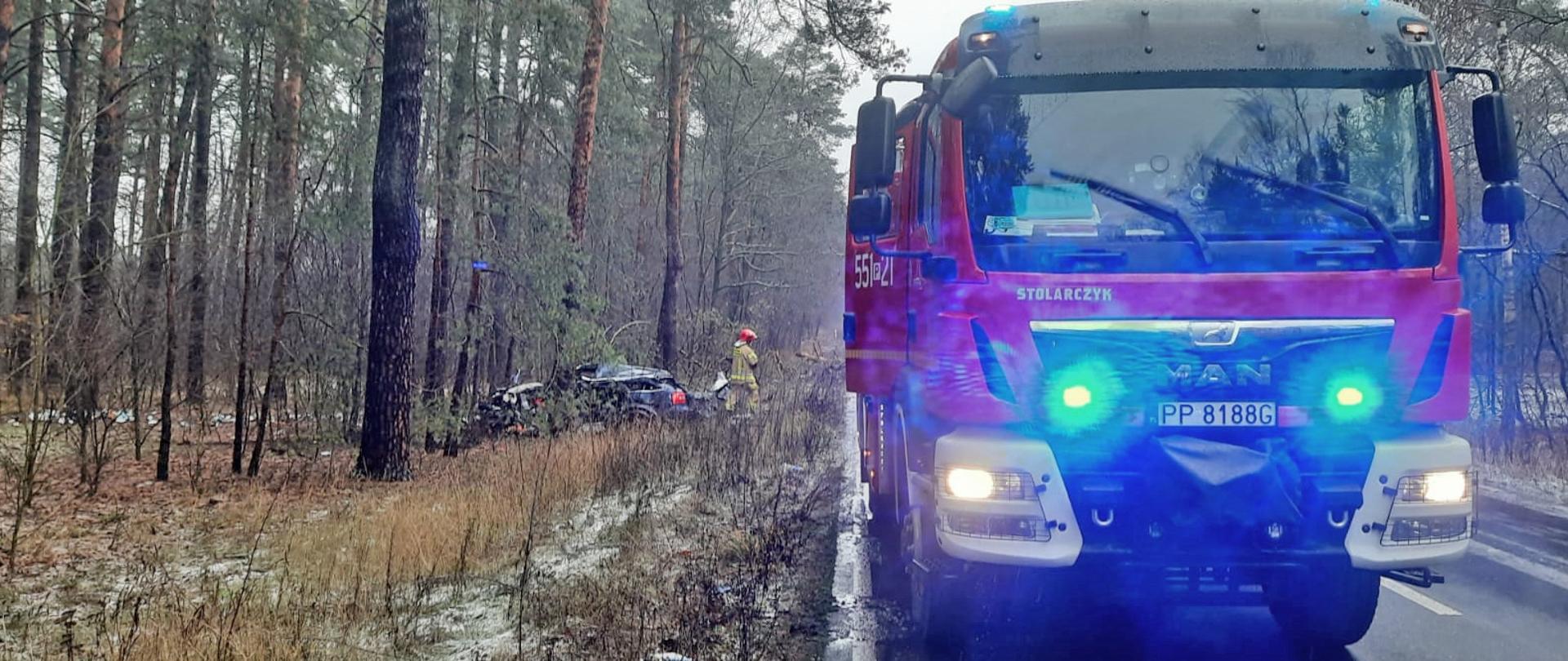 Na zdjęciu widać samochód pożarniczy na pierwszym planie. Dalej w lesie stoi uszkodzony pojazd a obok strażak.
