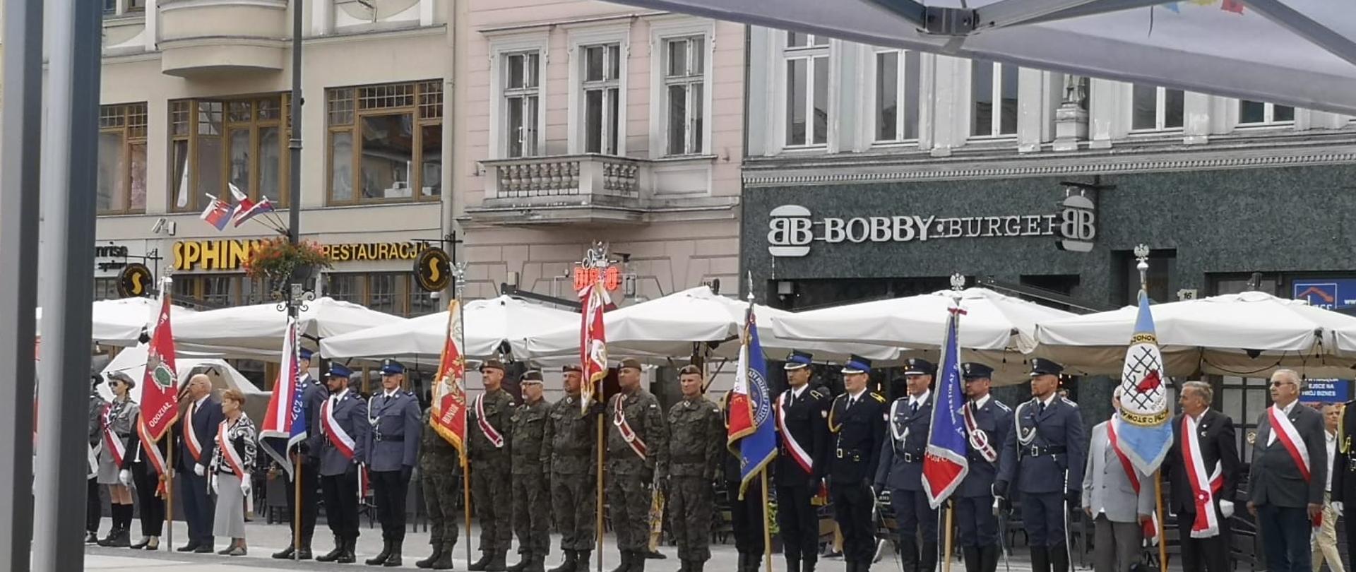 Poczty sztandarowe różnych służb stojące w szeregu na szarej płycie betonowej Starego Rynku w Bydgoszczy. W tle kamienice. 