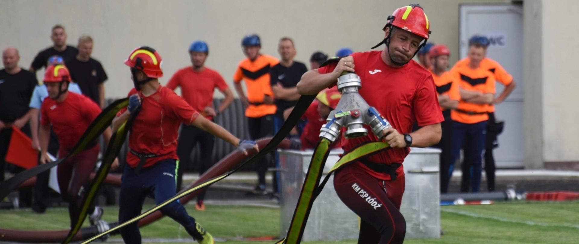 Na zdjęciu widać 3 strażaków w ubraniach sportowych wykonujących pożarnicze ćwiczenie bojowe w tle 9 osób i ściana