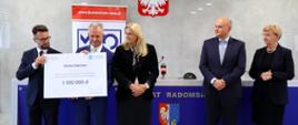 Pod szarą ścianą przed niebieskim stołem stoi pięć osób, dwaj mężczyźni z lewej trzymają wielki symboliczny czek z napisem Gmina Zakrzew - 1 000 000 zł.