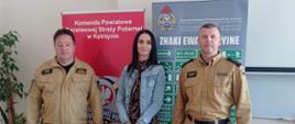 Zdjęcie przedstawiające trzy osoby: dwóch funkcjonariuszy straży pożarnej w mundurze służbowym koloru piaskowego stojących na po prawej i po lewej stronie oraz kobietę w ciemnej sukience z dżinsową kurtką stojącą po środku.