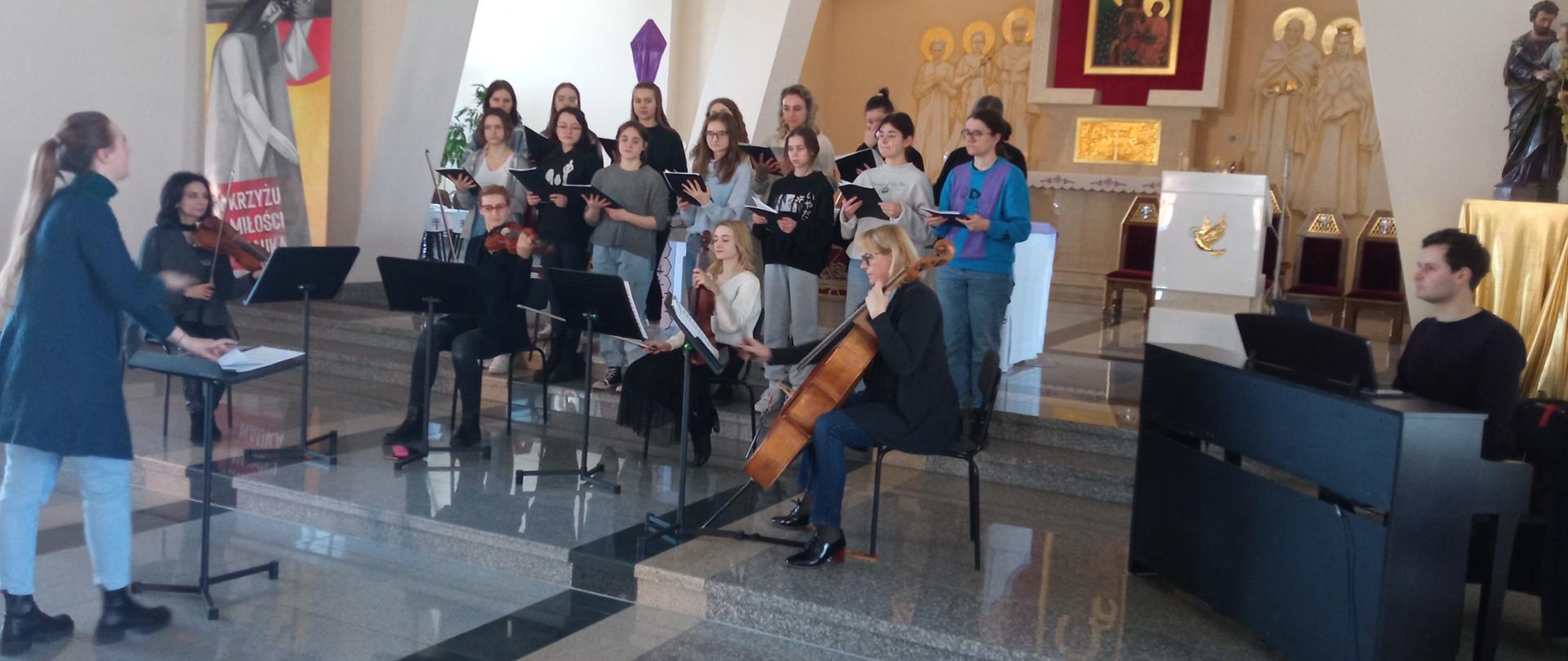 Zdjęcie z próby połączonych zespołów - instrumentalnego w postaci kwartetu smyczkowego wraz z gitarą i klawesynem oraz wokalnego w postaci Chóru Kameralnego ZPSM im. F. Chopina w Jarosławiu. Zdjęcie przedstawia ustawiony w świetle ołtarza w Kościół pw. Najświętszej Maryi Panny Królowej Polski w Jarosławiu chór kameralny, przed nim kwartet smyczkowy, po prawej stroni widoczny klawesynista realizujący partię na instrumencie elektronicznym. Przed kwartetem widoczna przy pulpicie dyrygenckim mgra Martyna Lasota