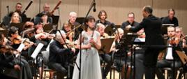 Zdjęcie przedstawiające flecistkę - Karolinę Laskowską na scenie filharmonii. W tle orkiestra symfoniczna. 