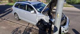 Zdjęcie pokazuje ten sam samochód, który uderzył przodem w metalowy słup oświetleniowy. Przednia część pojazdu jest poważnie uszkodzona. Maska jest uniesiona, a na słupie widnieje żółta tabliczka ostrzegawcza. W tle znajdują się domy mieszkalne i ogrodzenia. Ulica jest spokojna i nie widać innych pojazdów.