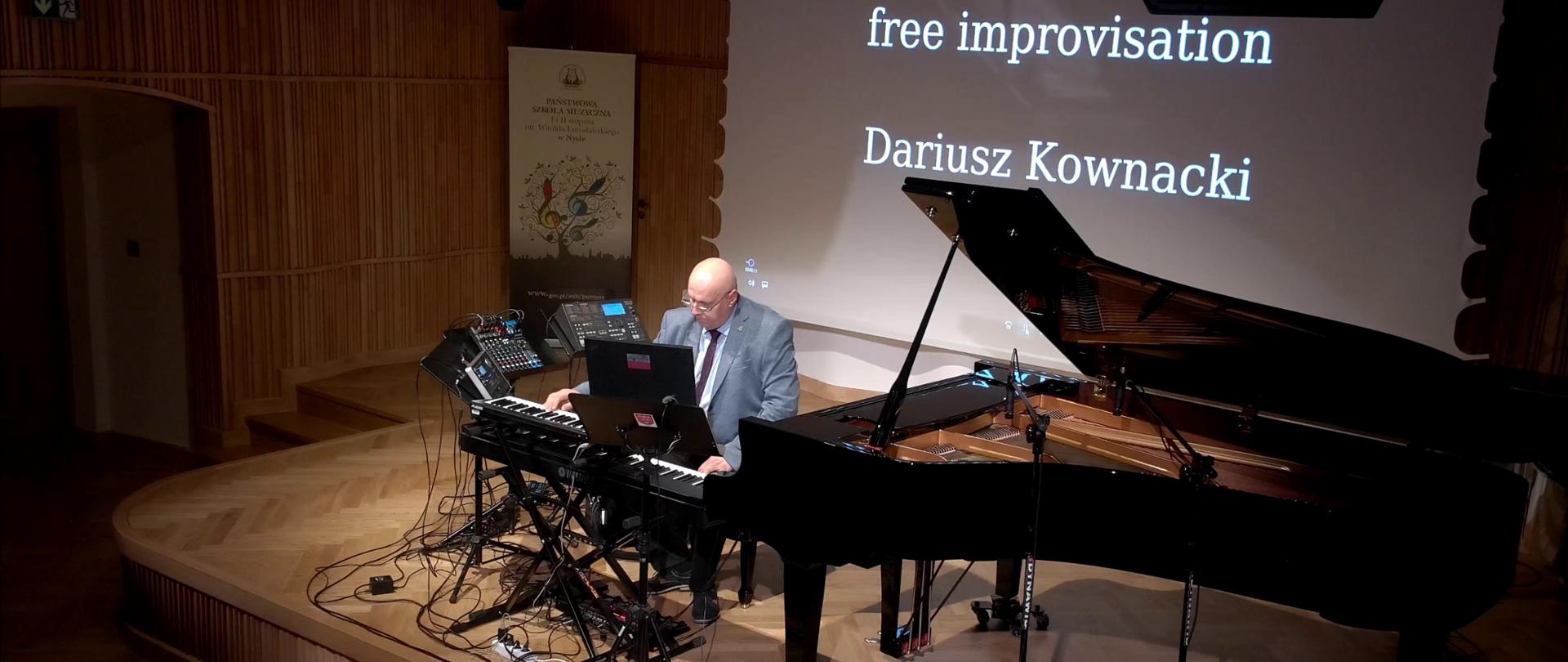Zdjęcie. Estrada sali koncertowej. Na estradzie otwarty duży czarny fortepian oraz zestaw oplecionych gęstwiną kabli instrumentów elektronicznych, na których gra pan Dariusz Kownacki. W tle na ekranie wyświetlony jest napis: free improvisation Dariusz Kownacki. W głębi w tle stoi baner PSM Nysa.