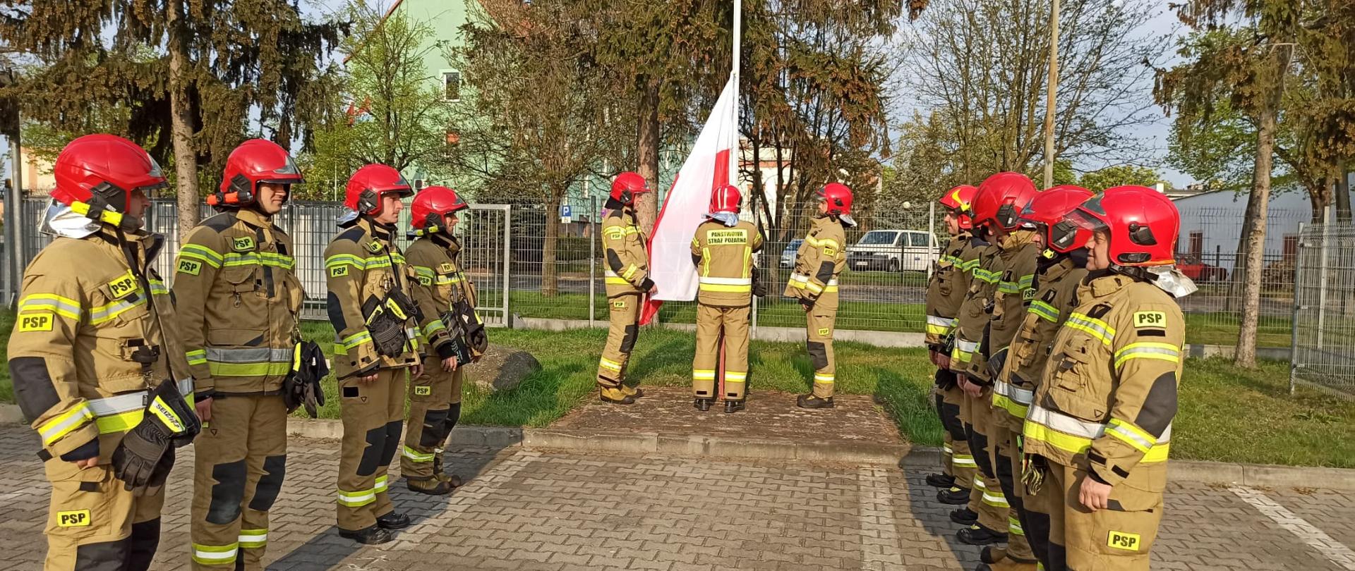 Trzech strażaków stojących z flagą przed masztem flagowych. W dwóch szeregach zwróceni twarzą do siebie stoją kolejni strażacy w umundurowaniu specjalnym.