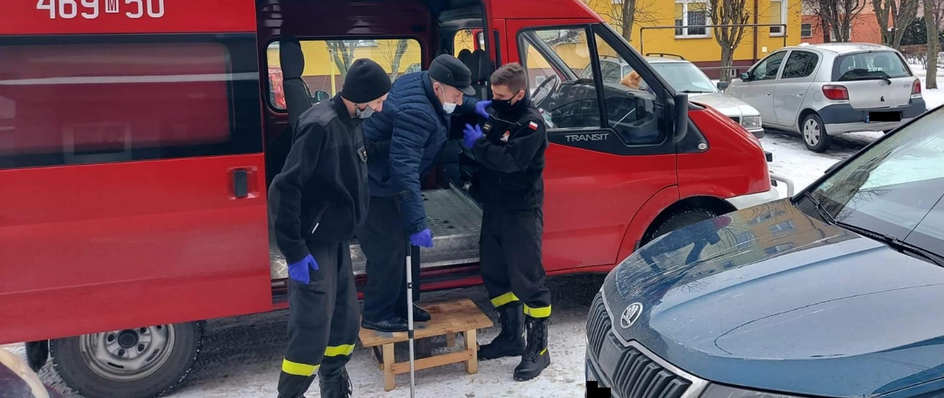 dwóch strażaków ochotniczej straży Pożarnej pomaga wysiąść z samochodu strażackiego seniorowi, który udaje się na szczepienie przeciw COVID-19