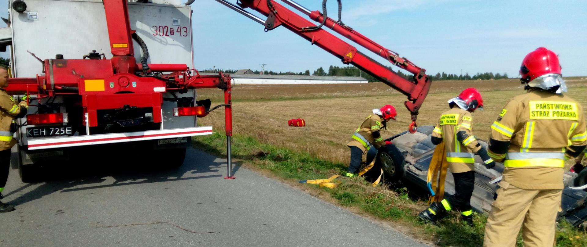 Zdjęcie przedstawia samochód osobowy, który leży na dachu w przydrożnym rowie. Strażacy podpinają taśmy na których auto zostanie ustawione na kołach.