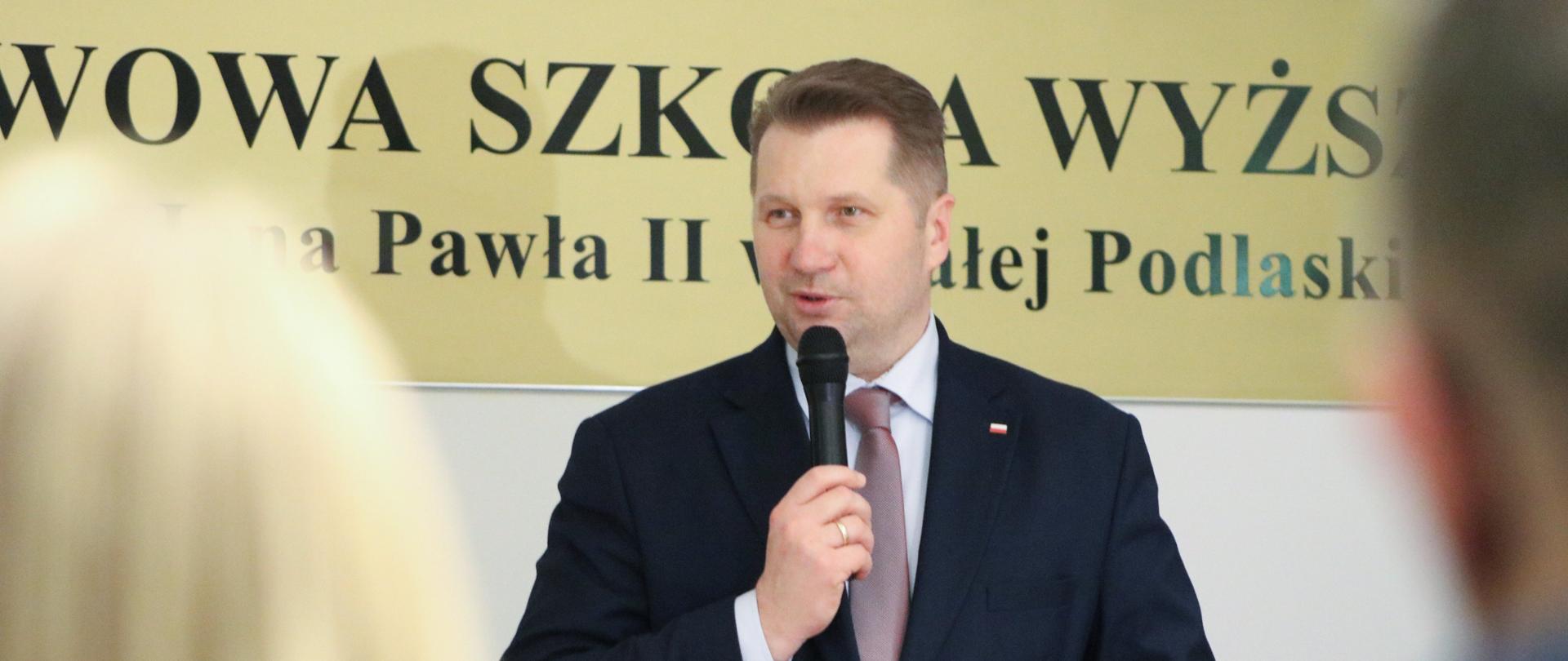 Minister Przemysław Czarnek przemawia podczas wystąpienia w wyższej szkole do mikrofonu. w tle nazwa uczelni. 