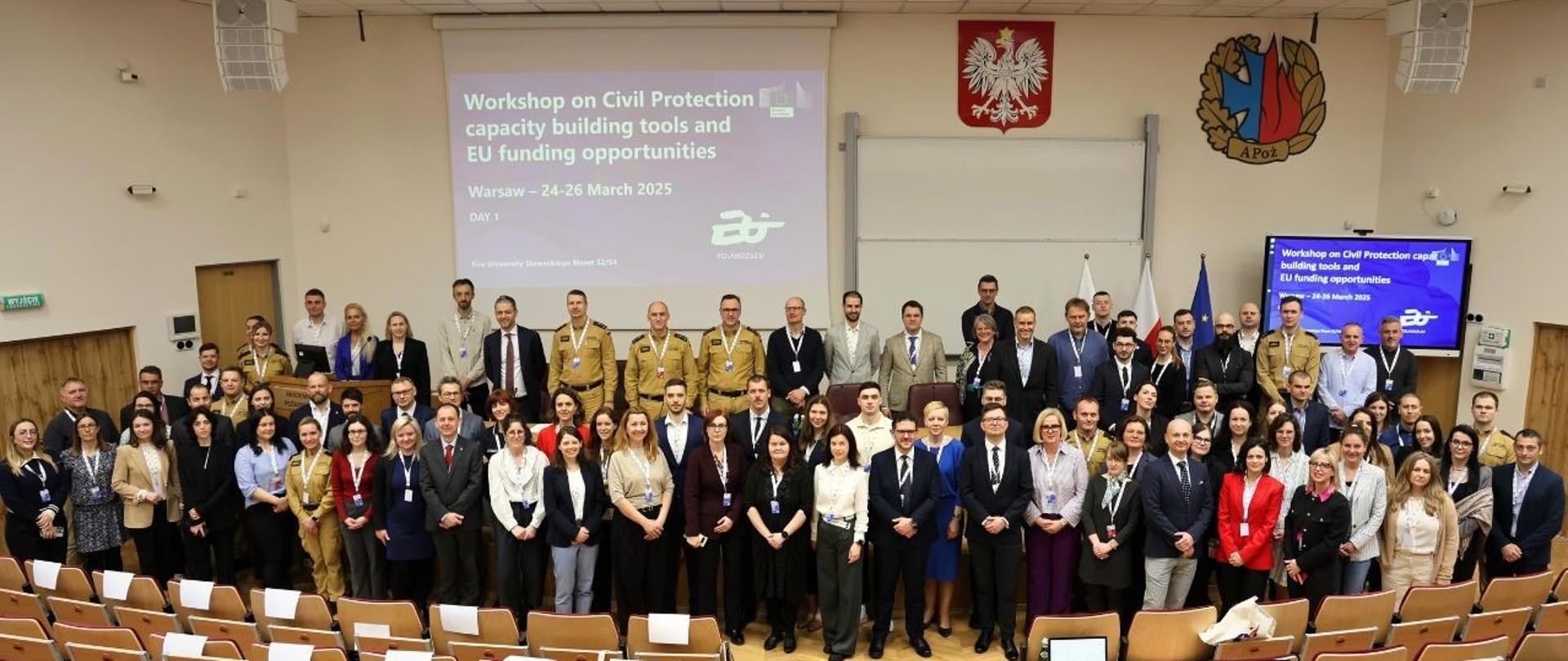 Zdjęcie grupowe wykonane w przestronnej auli wykładowej. Uczestnicy warsztatów stoją na tle dużego ekranu, na którym widnieje tytuł wydarzenia w języku angielskim: „Workshop on Civil Protection capacity building tools and EU funding opportunities, Warsaw – 24–26 March 2025”. Na ścianie nad tablicą znajdują się godło Polski i herb Akademii Pożarniczej. W pierwszych rzędach ustawiono kilkadziesiąt osób – kobiety i mężczyźni w strojach formalnych oraz kilku mężczyzn w piaskowych mundurach strażackich. W tle widać puste krzesła z pulpitami. Zdjęcie dokumentuje oficjalne rozpoczęcie wydarzenia o międzynarodowym charakterze.