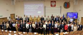 Zdjęcie grupowe wykonane w przestronnej auli wykładowej. Uczestnicy warsztatów stoją na tle dużego ekranu, na którym widnieje tytuł wydarzenia w języku angielskim: „Workshop on Civil Protection capacity building tools and EU funding opportunities, Warsaw – 24–26 March 2025”. Na ścianie nad tablicą znajdują się godło Polski i herb Akademii Pożarniczej. W pierwszych rzędach ustawiono kilkadziesiąt osób – kobiety i mężczyźni w strojach formalnych oraz kilku mężczyzn w piaskowych mundurach strażackich. W tle widać puste krzesła z pulpitami. Zdjęcie dokumentuje oficjalne rozpoczęcie wydarzenia o międzynarodowym charakterze.