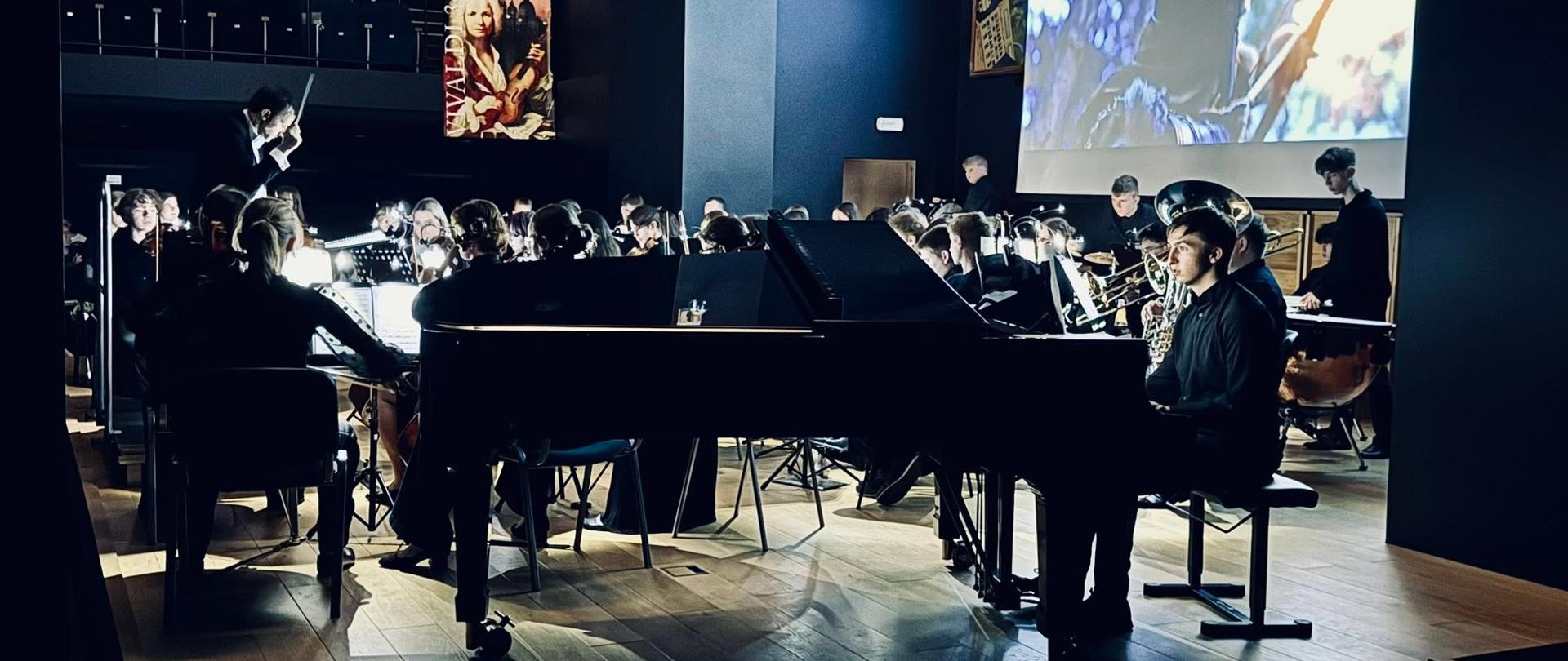 Koncert orkiestry smyczkowej w nowoczesnej sali koncertowej z okazji Dnia Otwartego (7.05). Na scenie młodzi muzycy grają na instrumentach smyczkowych - skrzypcach, altówkach, wiolonczelach i kontrabasach, pod kierunkiem dyrygenta. W tle wyświetlana jest prezentacja multimedialna z napisem "7.05 DZIEŃ OTWARTY" i ilustracją przedstawiającą dzieci grające na instrumentach. Publiczność siedzi na drewnianych ławkach i podłodze, obserwując występ. Sala ma nowoczesną architekturę z ciemnymi ścianami, profesjonalnym oświetleniem scenicznym i dużym ekranem projekcyjnym. 