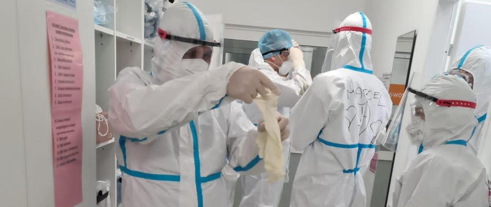 Zdjęcie przedstawia ratowników medycznych ubranych w białe, całościowe ubrania ochronne na tle pomieszczenia medycznego gdzie ratownicy ubierają się do czynności służbowych. Na pierwszym planie ratownik ubierający rękawice jednorazowe na dłonie
