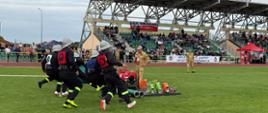 Strażacy w czarnych mundurach i jasnych hełmach dynamicznie wybiegają ze stanowiska ze sprzętem pożarniczym na płycie boiska pod nadzorem sędziego PSP podczas zawodów sportowo-pożarniczych.