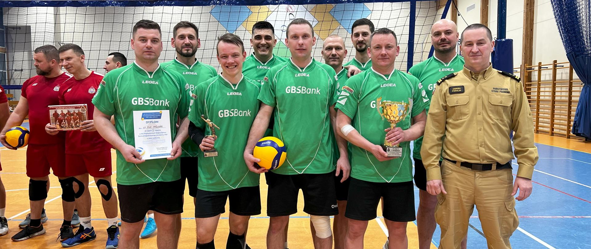 Zawodnicy zwycięskiej drużyny z komendy powiatowej Państwowej Straży Pożarnej z Myśliborza podczas pamiątkowego zdjęcia z zastępcą zachodniopomorskiego komendanta wojewódzkiego psp. Strażacy trzymają puchar, dyplom oraz piłkę. W tle widoczne jest boisko do gry w siatkówkę. Zdjęcie powiększy się po kliknięciu na nie.
