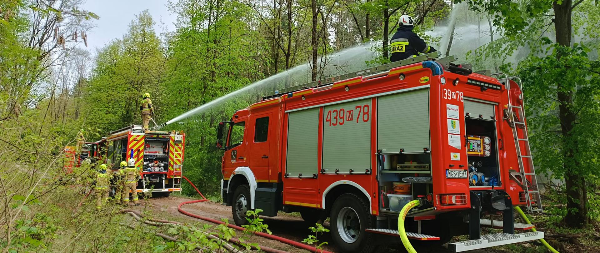 Czerwony wóz strażacki, z numerem "439 78" na boku, stoi na leśnej drodze. Strażak w żółto-czarnym mundurze i kasku, z napisem "STRAŻ" na plecach, stoi na dachu pojazdu, obsługując prądownicę, z której wydobywa się silny strumień wody. Przed wozem strażackim, w tle, widoczny jest kolejny wóz strażacki, z którego również wydobywa się strumień wody, obsługiwany przez innego strażaka. Obok tego drugiego wozu stoi kilku strażaków w pełnym umundurowaniu, a na ziemi widać węże strażackie. Scena rozgrywa się w lesie z zielonymi drzewami i jasnym niebem.