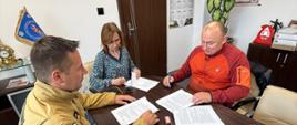 Podpisanie umowy w Komendzie Powiatowej PSP w Olecku – na zdjęciu trzech uczestników wydarzenia. Komendant PSP w mundurze, obok kobieta w sukience, mężczyzna w czerwonej bluzie, trzyma długopis.