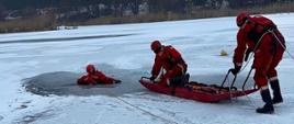 Ratownicy w czerwonych kombinezonach prowadzą akcję ratunkową na zamarzniętym jeziorze, wyciągając osobę z przerębla przy użyciu liny i sanek lodowych.