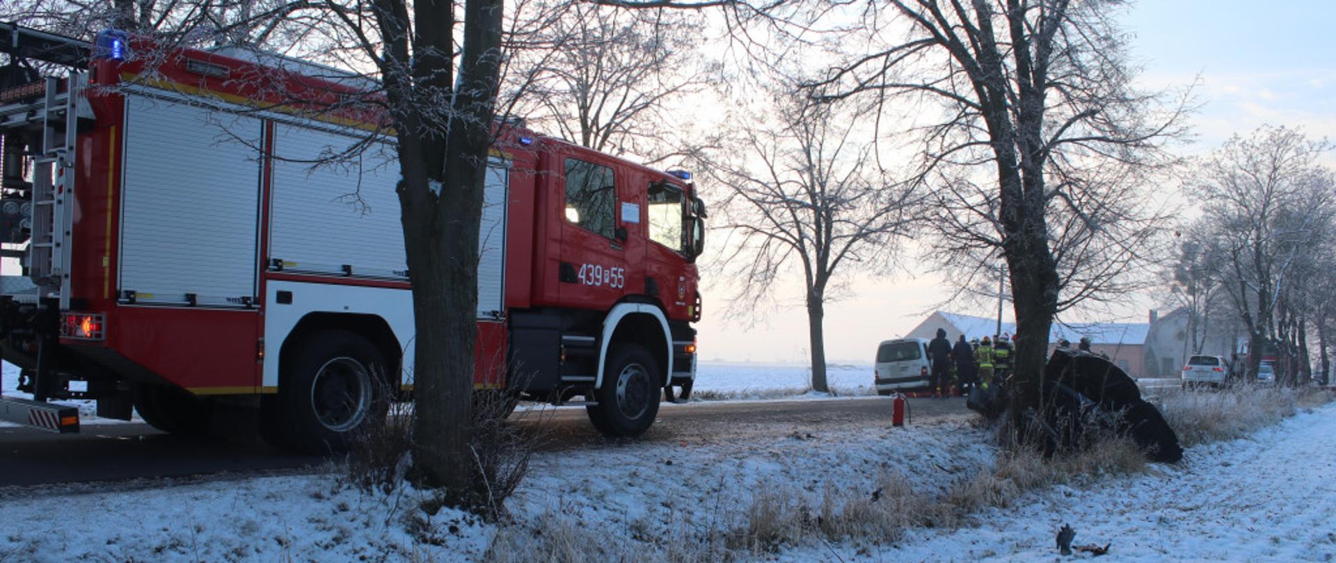 Zdjęcie przedstawia samochód pożarniczy. W tle znajduje się samochód osobowy po uderzeniu w drzewo.