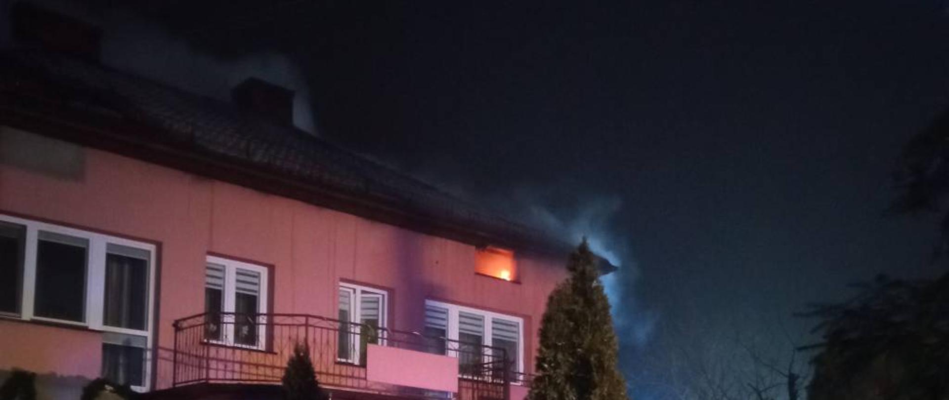 Zdjęcie przedstawia dom jednorodzinny, dwukondygnacyjny z poddasza którego wydobywa się szary dym. W jednym z okien na poddaszu widać płomienie. Zdjęcie wykonano w pogodną, zimową noc.