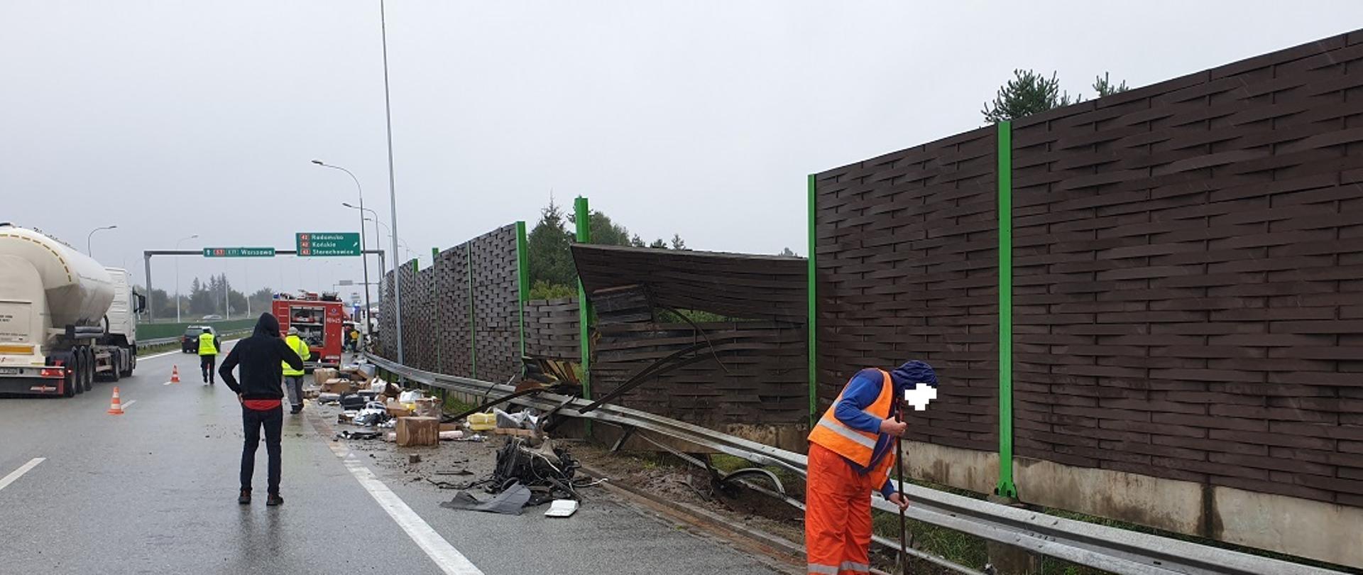 Na drodze szybkiego ruchu S-7 w miejscowości Skarżysko-Kamienna widoczna jest część pasa ruchu na którym doszło do wypadku drogowego. Z prawej strony widoczna jest uszkodzona bariera energochłonna, przy której widoczne są porozrzucane paczki przewożone przez pojazd uczestniczący w zdarzeniu oraz elementy karoserii tego pojazdu. Przy prawej krawędzi drogi widoczny jest pracownik służb drogowych wykonujący czynności polegające na usuwaniu skutków wypadku. W oddali widać funkcjonariusza policji oraz pracownika służb drogowych. Na drodze widoczna jest również osoba odwrócona tyłem rozmawiająca przez telefon komórkowy. Po lewej stronie stoi samochód ciężarowy typu cysterna. W oddali widać samochód straży pożarnej. 