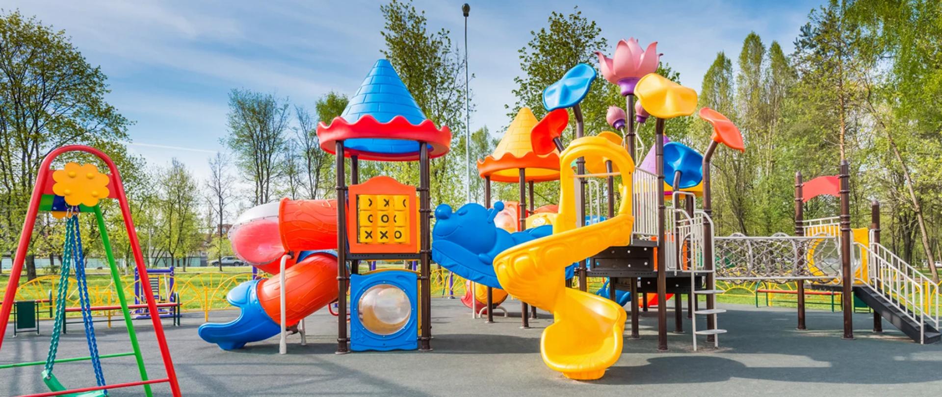 Na zdjęciu plac zabaw z kolorowymi zjeżdżalniami, huśtawkami, tunelami. W tle zielone drzewa. 