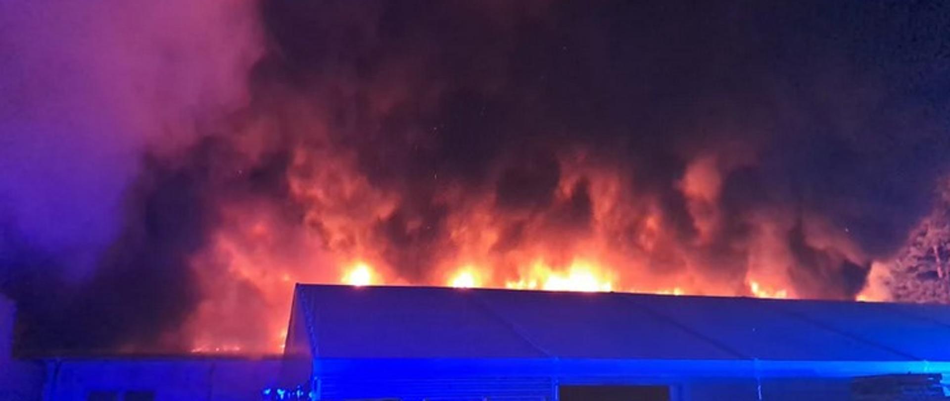 Zdjęcie zrobione w nocy. Na pierwszym planie hala z dachem dwuspadowym, za nim widać płomienie i dym palącego się innego budynku.