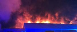 Zdjęcie zrobione w nocy. Na pierwszym planie hala z dachem dwuspadowym, za nim widać płomienie i dym palącego się innego budynku.