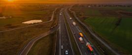 Widok z lotu ptaka na drogę ekspresową o zachodzie słońca; dwie jezdnie z ruchem pojazdów przebiegają przez tereny rolnicze i pola.