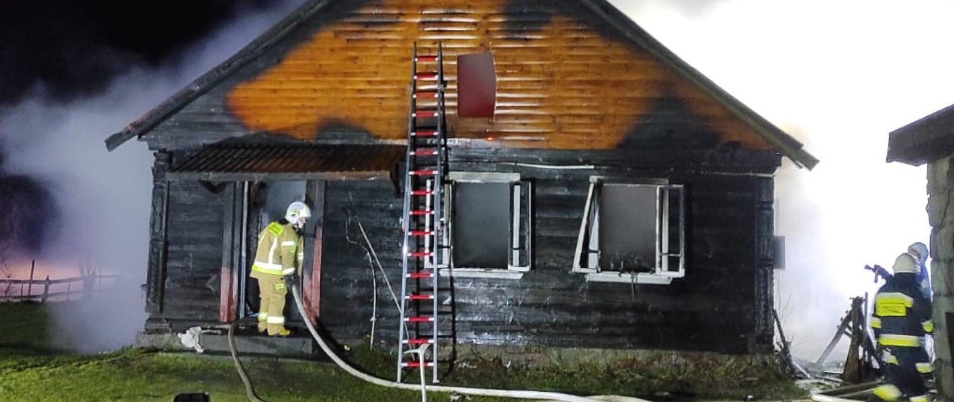 Budynek mieszkalny po pożarze. Konstrukcja ścian opalona. Nad budynkiem unosi się dym. Przy wejściu strażak prowadzący dogaszanie.