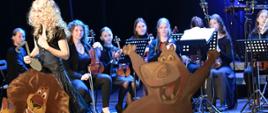 Z przodu dwie płaskie makiety postaci z bajek lwa i hipopotama, za nimi po lewej stronie kobieta w peruce blond mówi trzymając w ręce dużą nutę, z tyłu siedzi orkiestra.