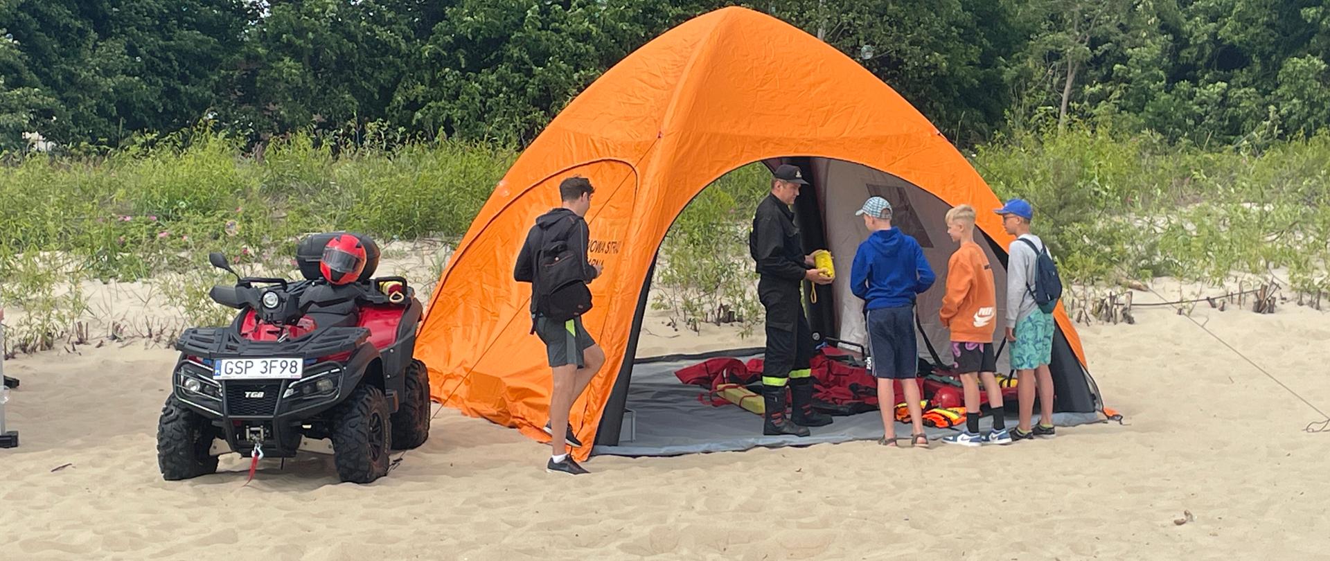 Sopocka plaża. Na niej w namiocie strażak ze sprzetem ratownictwa wodnego tłumaczy dzieciom zasady jego wykorzystania. Obok stoi quad